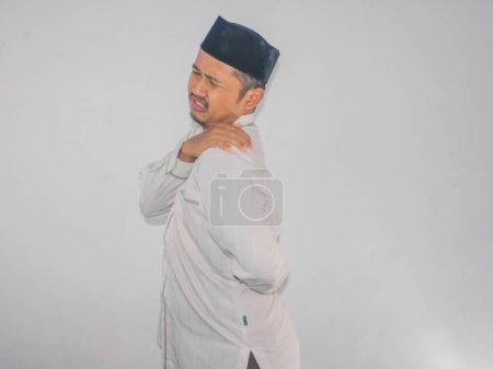 Asiatischer muslimischer Erwachsener berührt seine linke Schulter mit schmerzhaftem Gesichtsausdruck