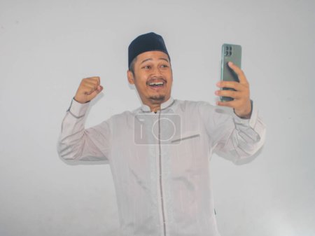 Téléchargez les photos : Musulman asiatique homme serré poing montrant l'excitation tout en regardant son téléphone mobile - en image libre de droit
