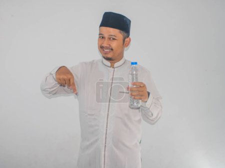 Moslem Asian man smiling and pointing finger down while holding a bottle of drinking water