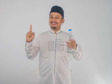 Hombre asiático musulmán sonriendo y señalando con el dedo hacia arriba mientras sostiene una botella de agua potable