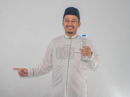 Téléchargez les photos : Homme musulman souriant tout en tenant une bouteille d'eau potable et pointant vers le côté droit - en image libre de droit