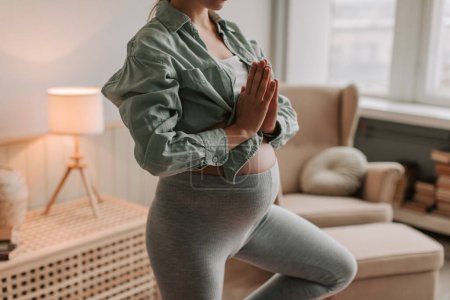 Nahaufnahme einer fitten Schwangeren in bequemer Kleidung beim Yoga-Balancieren auf einem Bein im heimischen Wohnzimmer. Menschen und Schwangerschaftsaktivitäten Konzept. Gesundes Elternkonzept. 