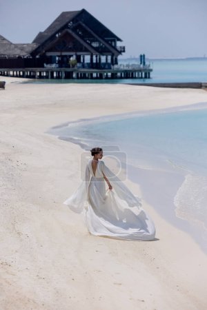 Téléchargez les photos : Jeune fille en robe blanche dansant sur le pont en bois aux Maldives. La bosse à l'arrière-plan. L'île. Photo de haute qualité - en image libre de droit