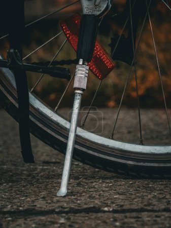 Photo for Close-up of a bicycle's rear wheel and kickstand on a textured pavement. The focus is on the metallic kickstand supporting the bike, with a clear view of the wheel, tire, and red reflector. The background is softly blurred - Royalty Free Image