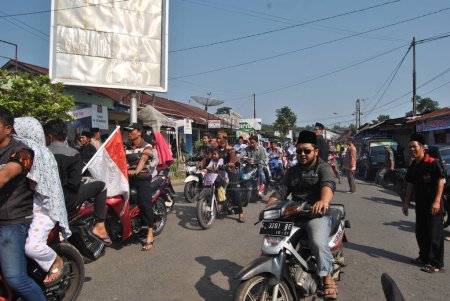 Téléchargez les photos : Tegal (INDONÉSIE), le 3 mai 2018 - Un groupe de gens du coin conduit des motos sans casque sur la route, suivant la troupe de carnaval devant eux - en image libre de droit