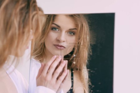 Photo for Beautiful caucasian blonde young woman in white shirt standing by the mirror. Health care, beauty, mental issues - Royalty Free Image