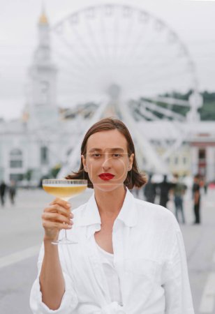 Photo for Beautiful woman is white dress is standing with the glass of wine on the city sqaure with the ferris wheel on the background. - Royalty Free Image