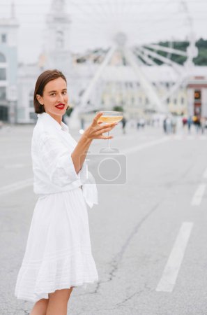 Téléchargez les photos : Belle femme est robe blanche est debout avec le verre de vin sur la place de la ville avec la roue ferris sur le fond. - en image libre de droit