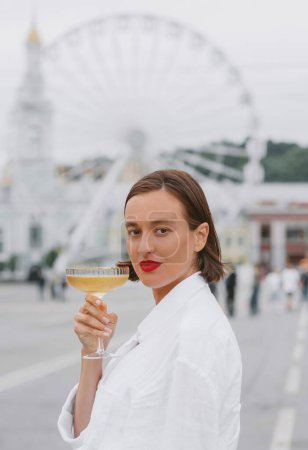 Photo for Beautiful woman is white dress is standing with the glass of wine on the city sqaure with the ferris wheel on the background. - Royalty Free Image