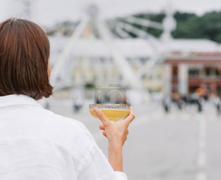 Téléchargez les photos : Dos de la femme est robe blanche debout avec le verre de vin sur la place de la ville avec la roue ferris sur le fond. - en image libre de droit