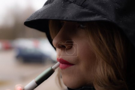 Photo for A woman with red lips is smoking an electronic cigarette. Beautiful young woman under the hood. Young girl with beautiful red lips - Royalty Free Image