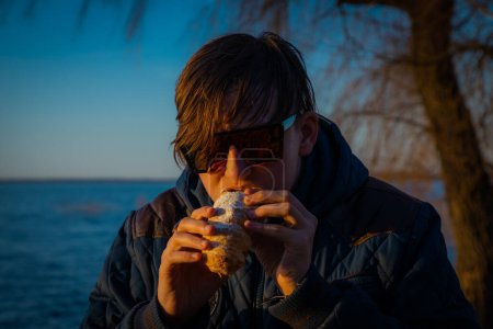 Photo for A boy in sunglasses watches the sunset. Young man wearing sunglasses enjoying summer. Lifestyle and summer. A man looks at the sun through sunglasses. The boy is eating the cake - Royalty Free Image