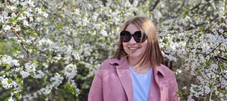 Foto de Mujer hermosa joven con gafas de sol abrigo rosa posando cerca de la acacia floreciente. Una chica con gafas de sol sonríe a la cámara. - Imagen libre de derechos