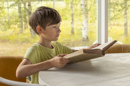 Téléchargez les photos : Un enfant s'assoit à la table et lit un vieux livre. Un garçon avec un livre à la main. Une leçon d'enfant avec un livre - en image libre de droit