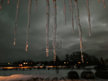 The serene winter landscape features glistening icicles hanging against a dusky sky. The snow-covered ground and trees are illuminated by distant lights. The overall atmosphere is chilly and calm.