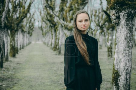 Frau in einer Lindenallee in dunklem kariertem Mantel mit langen Haaren vor verschwommenem natürlichen Hintergrund