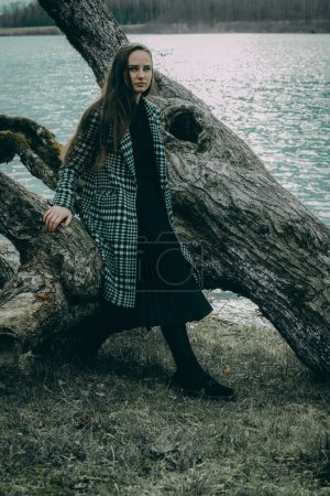 Eine Frau in einer Lindenallee in einem dunkel karierten Mantel mit langen Haaren vor verschwommenem natürlichen Hintergrund