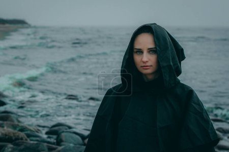 Eine geheimnisvolle weibliche Gestalt in schwarzer Kapuzenjacke steht am Strand vor verschwommenem Hintergrund, ihr Gesicht verborgen.