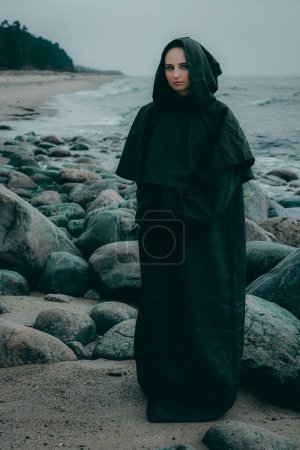 Foto de Una misteriosa figura femenina vestida con capucha negra se encuentra en la orilla del mar sobre un fondo borroso, su rostro oculto. - Imagen libre de derechos