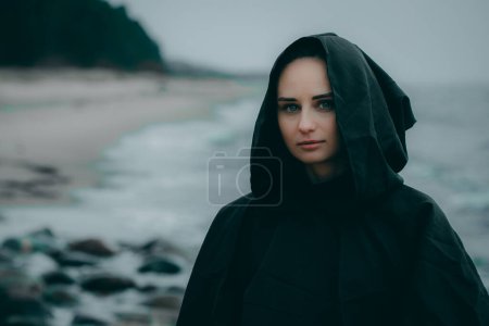 Foto de Una misteriosa figura femenina vestida con capucha negra se encuentra en la orilla del mar sobre un fondo borroso, su rostro oculto. - Imagen libre de derechos