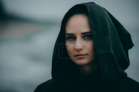 Photo for A mysterious female figure in black hooded clothing stands on the seashore against a blurred background, their face hidden. - Royalty Free Image