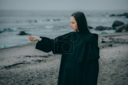 A mysterious female figure in black hooded clothing stands on the seashore against a blurred background, their face hidden.