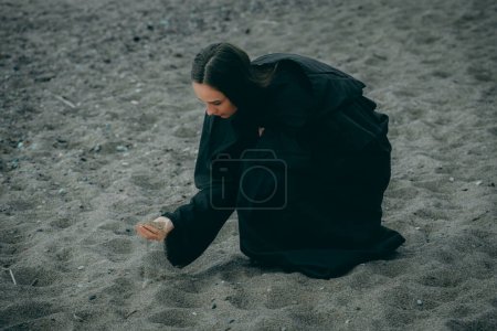 A woman by the sea in dark clothes takes sand in her hand on a blurred natural background.