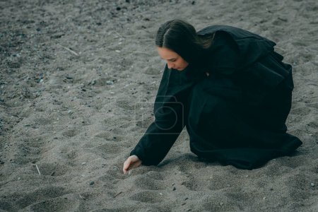 A woman by the sea in dark clothes takes sand in her hand on a blurred natural background.