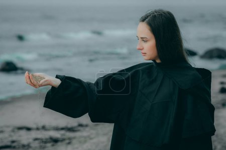 Frau in dunklem Kleid in der Nähe des Flusses hält Sand in der Hand, der auf einem verschwommenen natürlichen Hintergrund fällt.