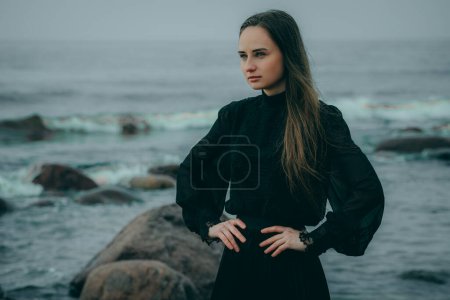 Frau am Meer in dunkler Kleidung mit langen Haaren vor verschwommenem natürlichen Hintergrund.