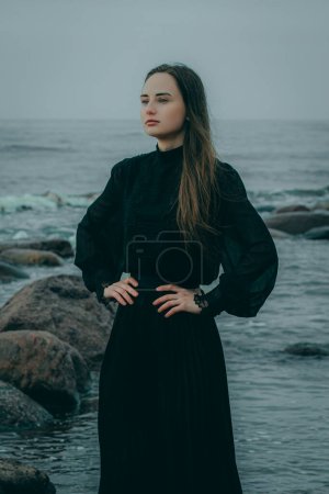 Frau am Meer in dunkler Kleidung mit langen Haaren vor verschwommenem natürlichen Hintergrund.