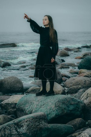 A silhouetted Woman stands by the rocky shore, pointing to the cloudy horizon. The waves crash against the rocks, creating a gloomy mood