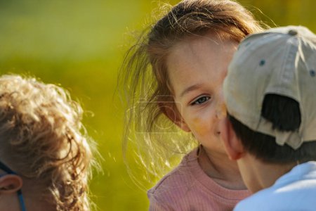 Téléchargez les photos : Une jeune fille chuchote à un ami dans un cadre extérieur ensoleillé, capturant un moment de secret et de connexion d'enfance. - en image libre de droit