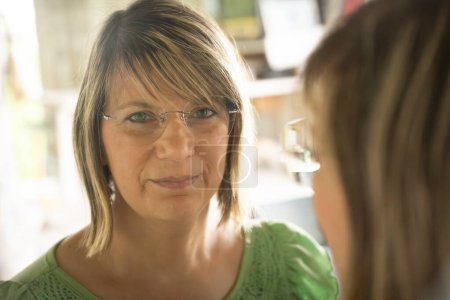Una mujer con gafas mira en un espejo, su reflejo parcialmente visible. Ella tiene el pelo rubio y lleva una parte superior verde, con iluminación interior suave creando un ambiente tranquilo e introspectivo.