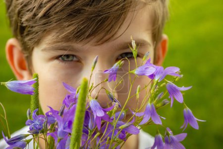 a boy with brown hair and blue eyes peeking through a bouquet of purple flowers. The image captures a moment of curiosity and playfulness with a bright green background.