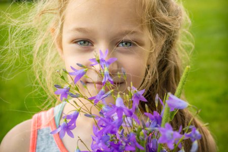 a child with blue eyes and blonde hair peeking through purple flowers, smiling softly. The image captures a moment of innocence and joy with natural lighting.
