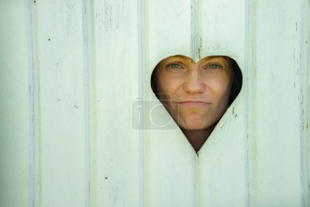 A woman looks through a heart-shaped cutout in a rustic white door, giving a humorous expression. The image captures a candid moment with an outdoor setting.