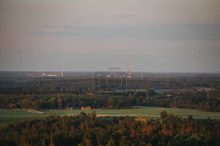 A panoramic view of lush forest and farmland at sunset, with a distant industrial skyline featuring tall towers. The warm light casts a serene glow over the landscape