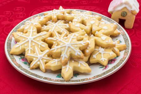 Plate with snowflake-shaped sugar icing Christmas cookies. Disposed on a red tablecloth with a house-shaped cookie as decoration.