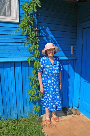 Téléchargez les photos : Femme âgée en chapeau sur le porche du bâtiment rural en bois - en image libre de droit