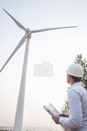 Téléchargez les photos : Jeune ingénieur en casque rigide regardant une éolienne, plan à la main - en image libre de droit
