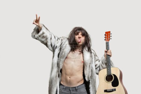 Téléchargez les photos : Jeune homme en manteau de fourrure tenant la guitare tout en gesticulant sur fond gris - en image libre de droit