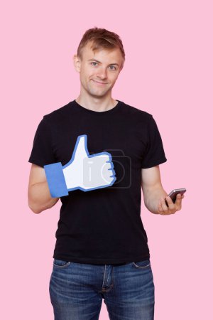 Photo for "Portrait of a happy young man with cell phone holding fake like button over pink background" - Royalty Free Image
