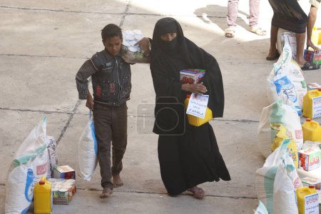 Foto de YEMEN, Taiz: Una mujer yemení y su hijo llevan bolsas de suministros durante una distribución de alimentos y agua limpia en la escuela Nasser, en las afueras de Taiz (Taiz), al oeste de Yemen, el 23 de octubre de 2015.. - Imagen libre de derechos
