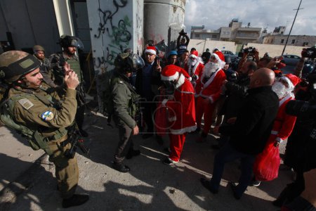 Téléchargez les photos : BANQUE DE L'OUEST, Bethléem : Des manifestants palestiniens en costumes de Père Noël agitent le drapeau palestinien lors d'une marche de protestation à Bethléem le 18 décembre 2015, alors que les soldats israéliens regardent. - en image libre de droit