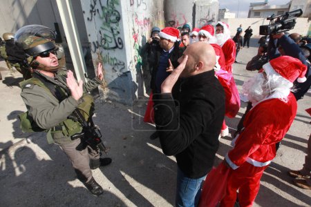 Téléchargez les photos : BANQUE DE L'OUEST, Bethléem : Des manifestants palestiniens en costumes de Père Noël agitent le drapeau palestinien lors d'une marche de protestation à Bethléem le 18 décembre 2015, alors que les soldats israéliens regardent. - en image libre de droit