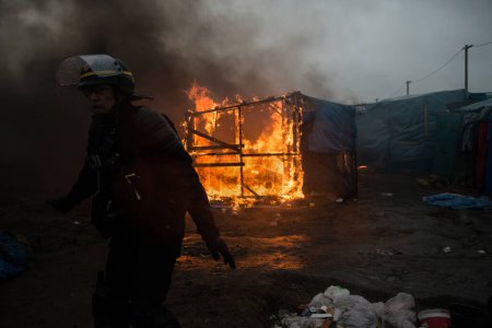 Foto de FRANCIA, Calais: Los migrantes pasan por una chozas incendiarias, en la parte sur del llamado campamento de migrantes de la Selva, mientras la mitad del campamento está siendo desmantelado, en la ciudad portuaria francesa del norte de Calais, el 1 de marzo de 2016 - Imagen libre de derechos