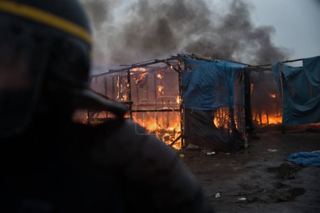 Foto de FRANCIA, Calais: Los migrantes pasan por una chozas incendiarias, en la parte sur del llamado campamento de migrantes de la Selva, mientras la mitad del campamento está siendo desmantelado, en la ciudad portuaria francesa del norte de Calais, el 1 de marzo de 2016 - Imagen libre de derechos