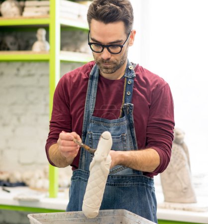 Téléchargez les photos : Portrait de céramiste vêtu d'un tablier travaillant sur la sculpture en argile dans un atelier de céramique lumineuse. - en image libre de droit