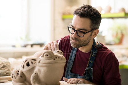 Téléchargez les photos : Portrait de céramiste vêtu d'un tablier travaillant sur la sculpture en argile dans un atelier de céramique lumineuse. - en image libre de droit